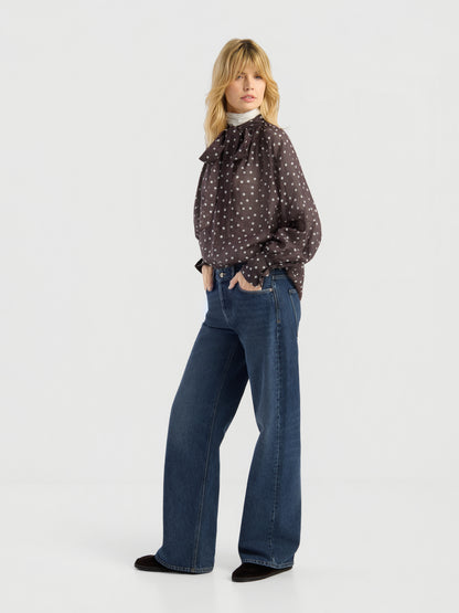 Vor einem weißen Hintergrund steht eine Frau mit blondem, welligem Haar. Sie trägt die Bluse „Dotty Bow Brown Ecru“ zu einer blauen Hose mit weitem Bein und schwarzen Schuhen. Ihre Hände hat sie in die Hosentaschen gesteckt.