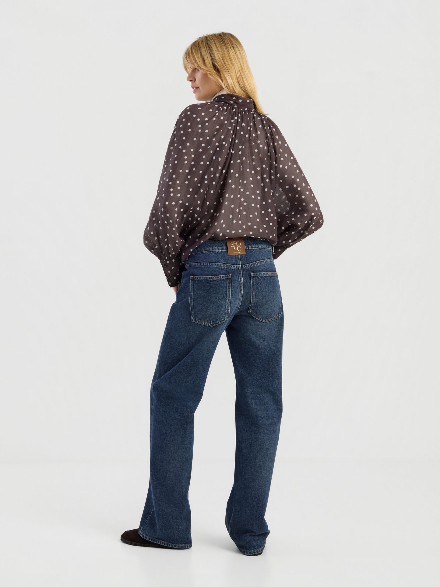 Eine Person mit blonden Haaren steht mit dem Rücken zur Kamera und trägt die „Blouse Dotty Bow Brown Ecru“ – eine locker sitzende braune Bluse mit weißen Punkten – sowie eine blaue Jeans mit hoher Taille und einem braunen Lederaufnäher über der rechten Tasche, vor dem Hintergrund eines minimalistisch gestalteten weißen Ateliers.