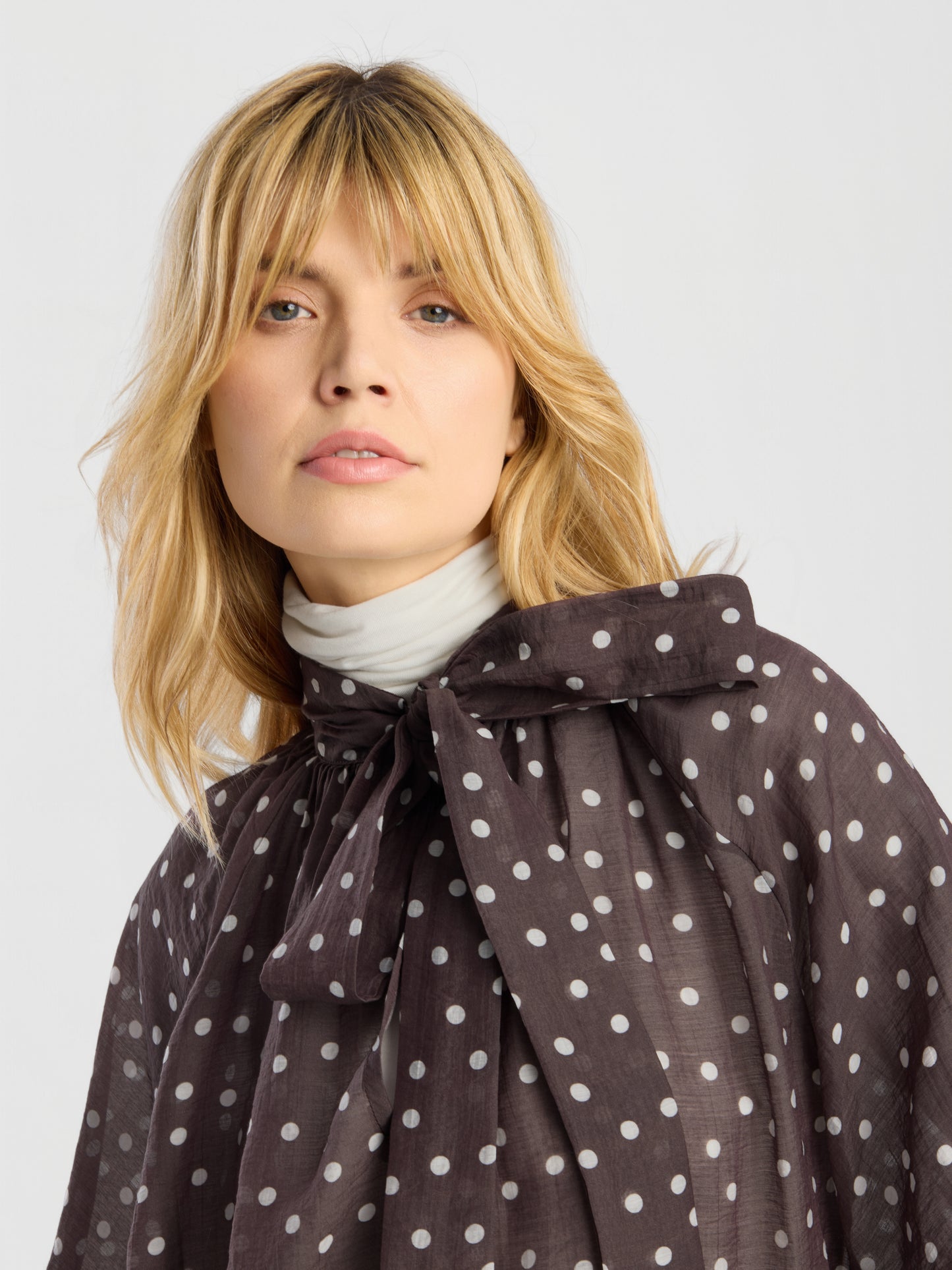 Eine blonde Frau mit Pony trägt die Bluse „Dotty Bow Brown Ecru“ mit Polka-Dots und einer großen Schleife über einem weißen Rollkragenpullover. Vor einem hellen Hintergrund blickt sie leicht an der Kamera vorbei.