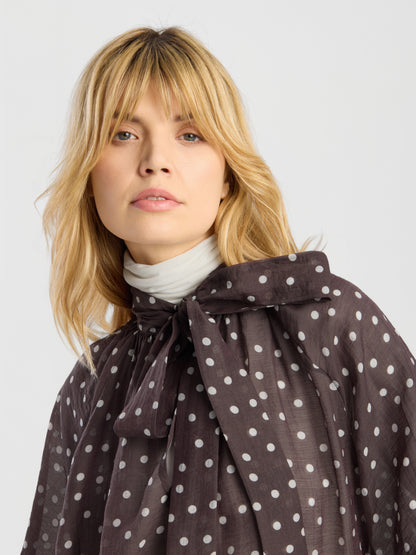 Eine blonde Frau mit Pony trägt die Bluse „Dotty Bow Brown Ecru“ mit Polka-Dots und einer großen Schleife über einem weißen Rollkragenpullover. Vor einem hellen Hintergrund blickt sie leicht an der Kamera vorbei.