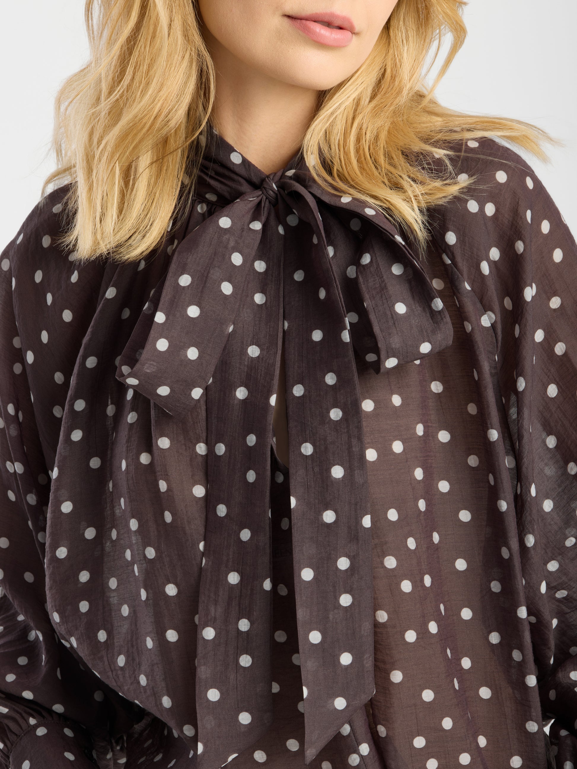 Eine blonde Frau trägt die Bluse „Dotty Bow Brown Ecru“, eine schicke Damenbluse mit weißen Polka-Punkten und einer großen Schleife am Ausschnitt, gefertigt aus luftigem Baumwoll-Voile.