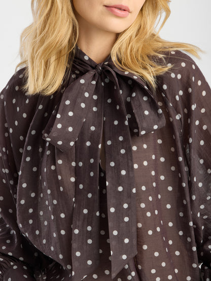Eine blonde Frau trägt die Bluse „Dotty Bow Brown Ecru“, eine schicke Damenbluse mit weißen Polka-Punkten und einer großen Schleife am Ausschnitt, gefertigt aus luftigem Baumwoll-Voile.