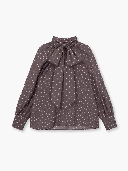 Die Bluse „Dotty Bow Grey Latte“ ist eine langärmelige Bluse aus braunem Baumwollvoile mit weißem Polka-Dot-Muster, hohem Kragen und einer übergroßen Schleife am Kragen. Sie wird in Italien hergestellt und ist hier vor einem schlichten weißen Hintergrund abgebildet.