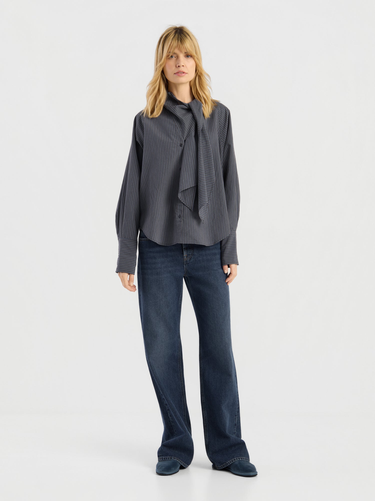 Vor einem schlichten Hintergrund steht eine Frau, die eine locker sitzende, dunkelgraue Bluse mit Streifenmuster, eine blaue Jeans mit weitem Bein und dunkle Schuhe trägt. Diese elegante Damenbluse „Made in Italy“ verleiht ihrem modernen Look eine raffinierte Note.