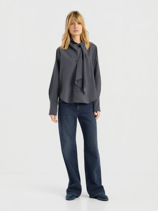 Eine Frau steht mit dem Blick nach vorne vor einem schlichten weißen Hintergrund. Sie trägt die dunkel gestreifte Bluse „Modern Stripes“ mit Krawatte, dazu eine blaue Jeans mit weitem Bein und dunkle Schuhe.