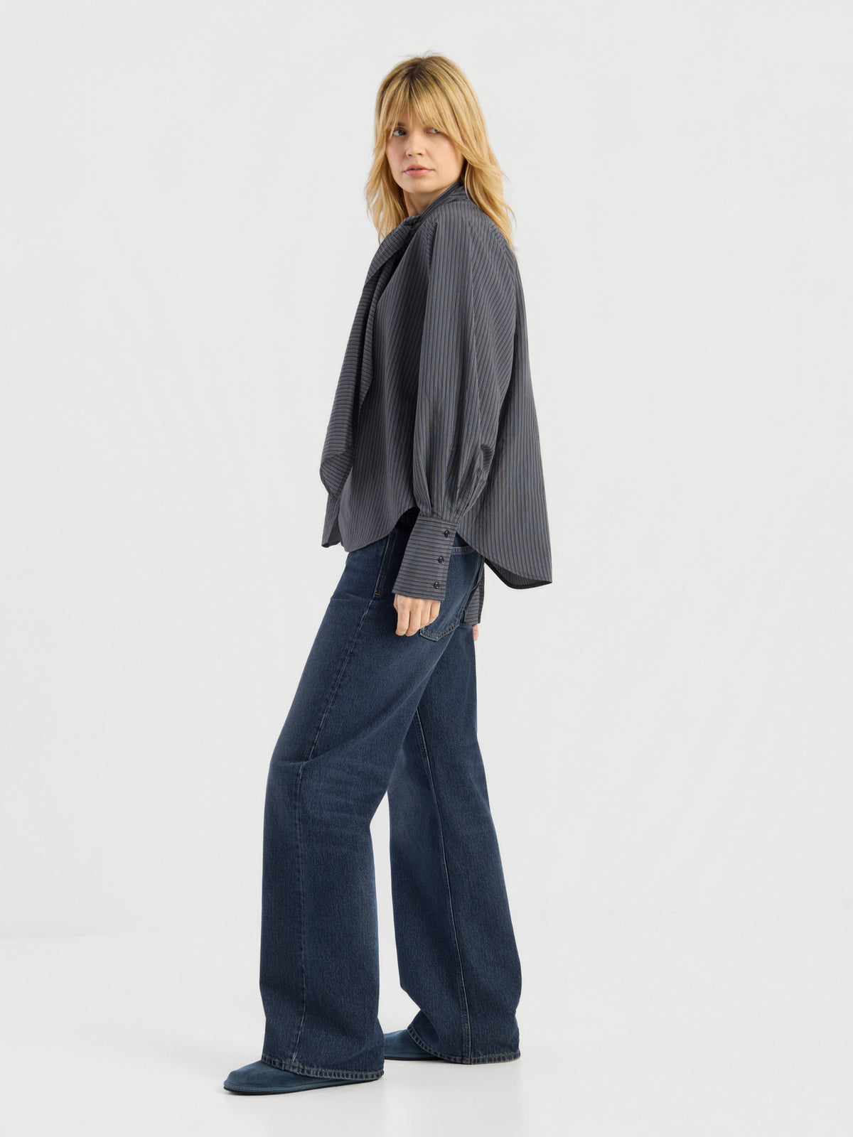 Eine Person mit blonden Haaren trägt einen locker sitzenden, dunklen Bluse Scarf Stripes, kombiniert mit einer blau-weit geschnittenen Jeans. Sie steht seitlich und blickt vor einem schlichten weißen Hintergrund in die Kamera. Dieses elegante Kleidungsstück wurde in Italien hergestellt und besticht sowohl durch seinen Stil als auch durch seine hochwertige Verarbeitung.