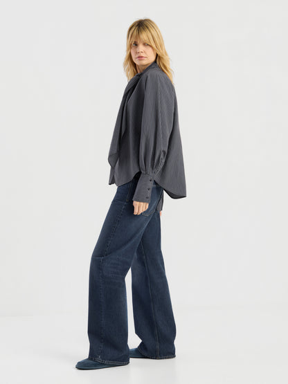 Eine Person mit blonden Haaren trägt einen locker sitzenden, dunklen Bluse Scarf Stripes, kombiniert mit einer blau-weit geschnittenen Jeans. Sie steht seitlich und blickt vor einem schlichten weißen Hintergrund in die Kamera. Dieses elegante Kleidungsstück wurde in Italien hergestellt und besticht sowohl durch seinen Stil als auch durch seine hochwertige Verarbeitung.