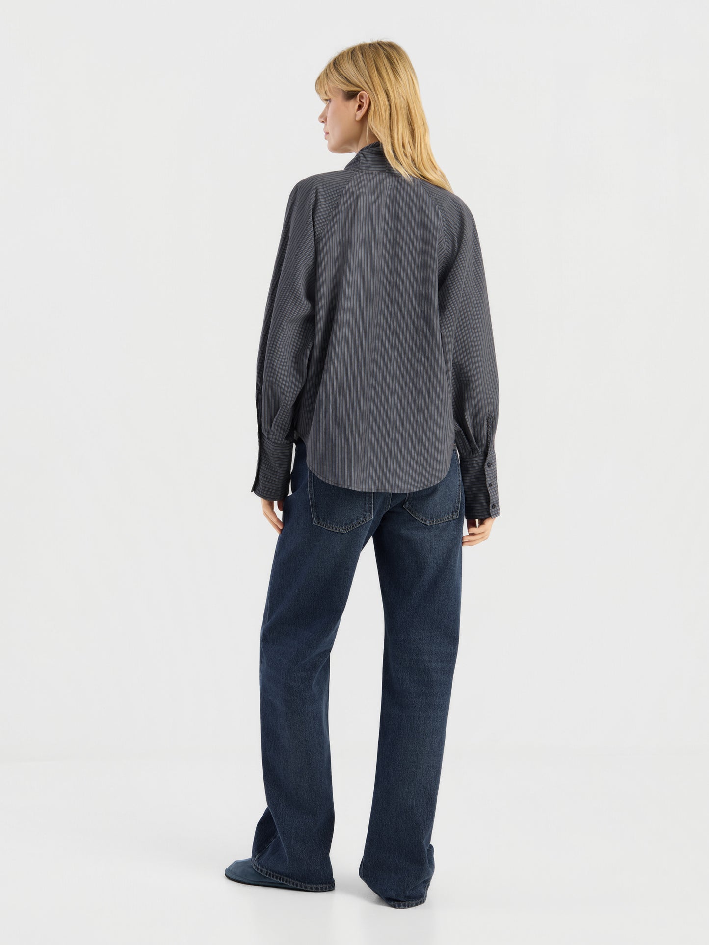 Vor einem schlichten weißen Hintergrund steht eine Person mit blonden Haaren mit dem Rücken zur Kamera. Sie trägt eine lockere, dunkelgraue Bluse mit Streifenmuster und langen Ärmeln sowie dunkelblaue Jeans. Diese stilvolle Damenbluse wurde in Italien hergestellt.