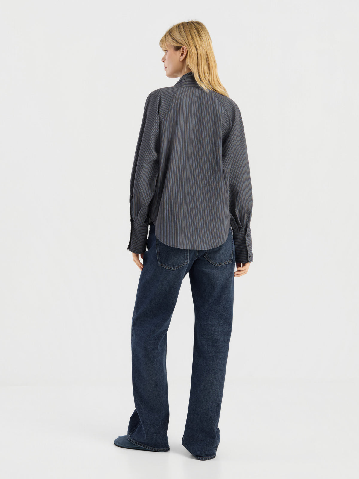 Vor einem schlichten weißen Hintergrund steht eine Person mit blonden Haaren mit dem Rücken zur Kamera. Sie trägt eine lockere, dunkelgraue Bluse mit Streifenmuster und langen Ärmeln sowie dunkelblaue Jeans. Diese stilvolle Damenbluse wurde in Italien hergestellt.