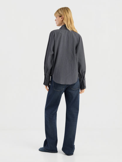 Vor einem schlichten weißen Hintergrund steht eine Person mit blonden Haaren mit dem Rücken zur Kamera. Sie trägt eine lockere, dunkelgraue Bluse mit Streifenmuster und langen Ärmeln sowie dunkelblaue Jeans. Diese stilvolle Damenbluse wurde in Italien hergestellt.