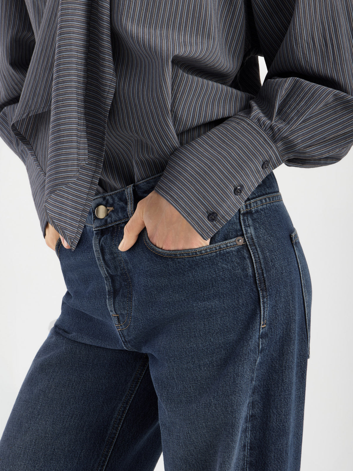 Diese in Italien gefertigte Damenbluse mit dem Modell „Bluse Scarf Stripes“ – einem dunklen Streifenmuster und geknöpften Manschetten – wird in Kombination mit einer blauen Jeans und den Händen in den Vordertaschen auf einem Bild gezeigt, das von den Schultern bis zur Mitte des Oberschenkelsausschnitt ist.