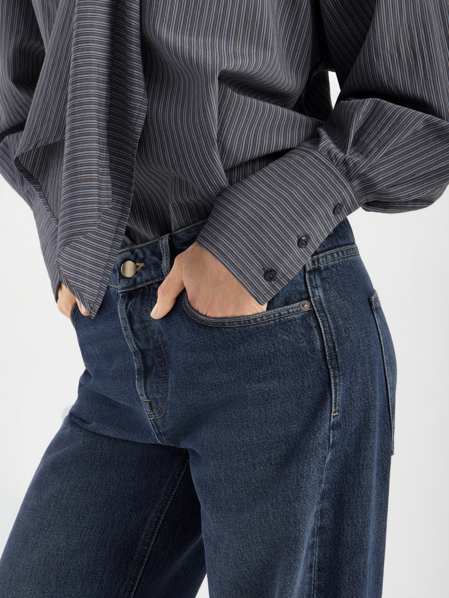 Diese in Italien gefertigte Damenbluse mit dem Modell „Bluse Scarf Stripes“ – einem dunklen Streifenmuster und geknöpften Manschetten – wird in Kombination mit einer blauen Jeans und den Händen in den Vordertaschen auf einem Bild gezeigt, das von den Schultern bis zur Mitte des Oberschenkelsausschnitt ist.