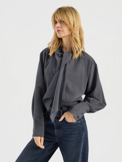 Eine Frau mit blonden Haaren trägt eine graue Bluse mit Streifenmuster und eine blaue Jeans. Sie steht mit einer Hand in der Tasche vor einem schlichten, hellen Hintergrund und präsentiert diese elegante, in Italien hergestellte Damenbluse.