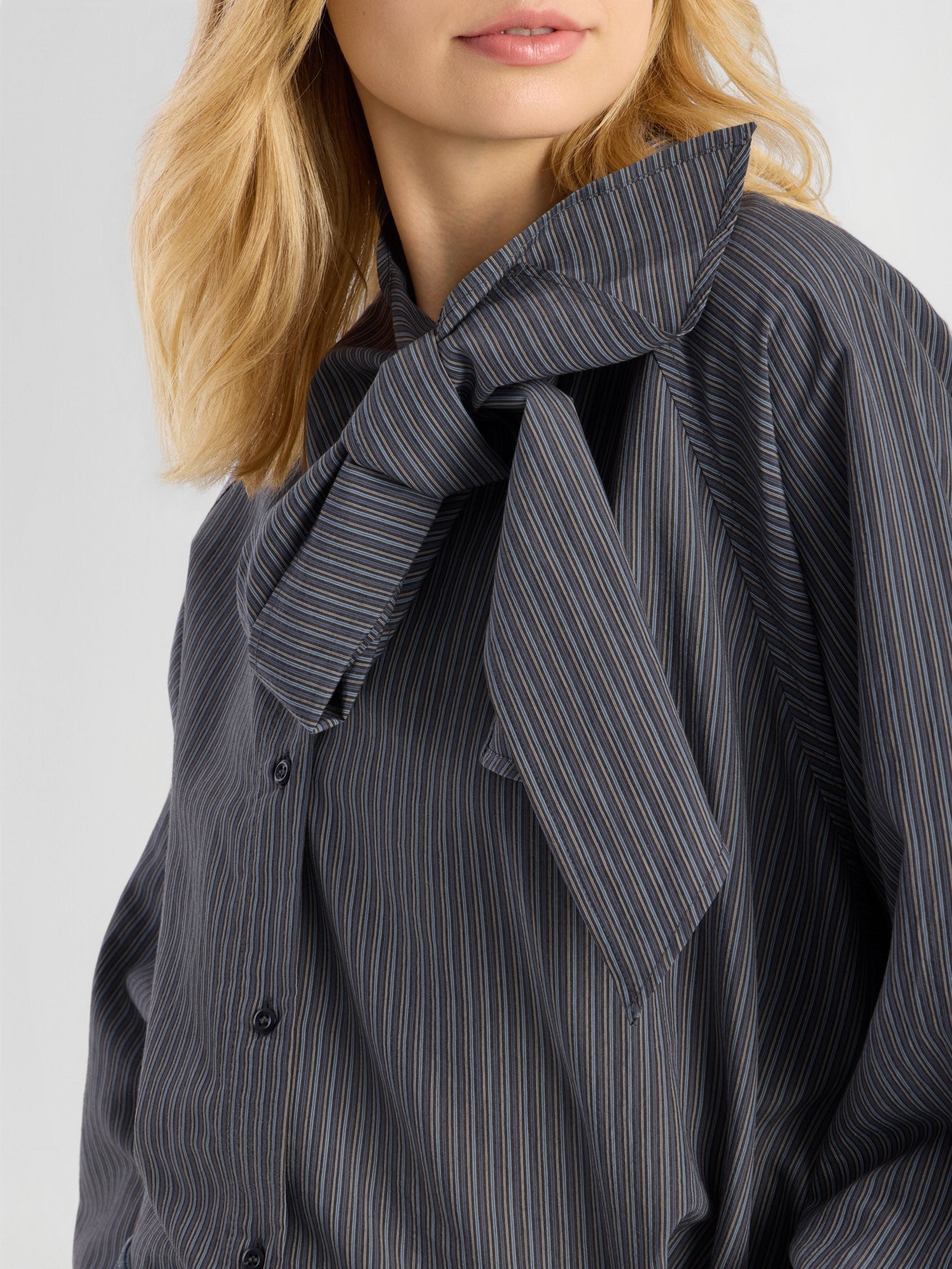 Eine Frau mit blonden Haaren trägt die dunkle Bluse „Scarf Stripes“, die sich durch eine auffällige Schleife am Kragen auszeichnet. Nur die untere Hälfte ihres Gesichts und der obere Oberkörper sind zu sehen, wodurch die elegante, in Italien gefertigte Damenbluse besonders gut zur Geltung kommt.