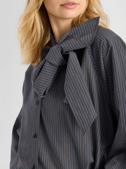 Eine Frau mit blonden Haaren trägt die dunkle Bluse „Scarf Stripes“, die sich durch eine auffällige Schleife am Kragen auszeichnet. Nur die untere Hälfte ihres Gesichts und der obere Oberkörper sind zu sehen, wodurch die elegante, in Italien gefertigte Damenbluse besonders gut zur Geltung kommt.