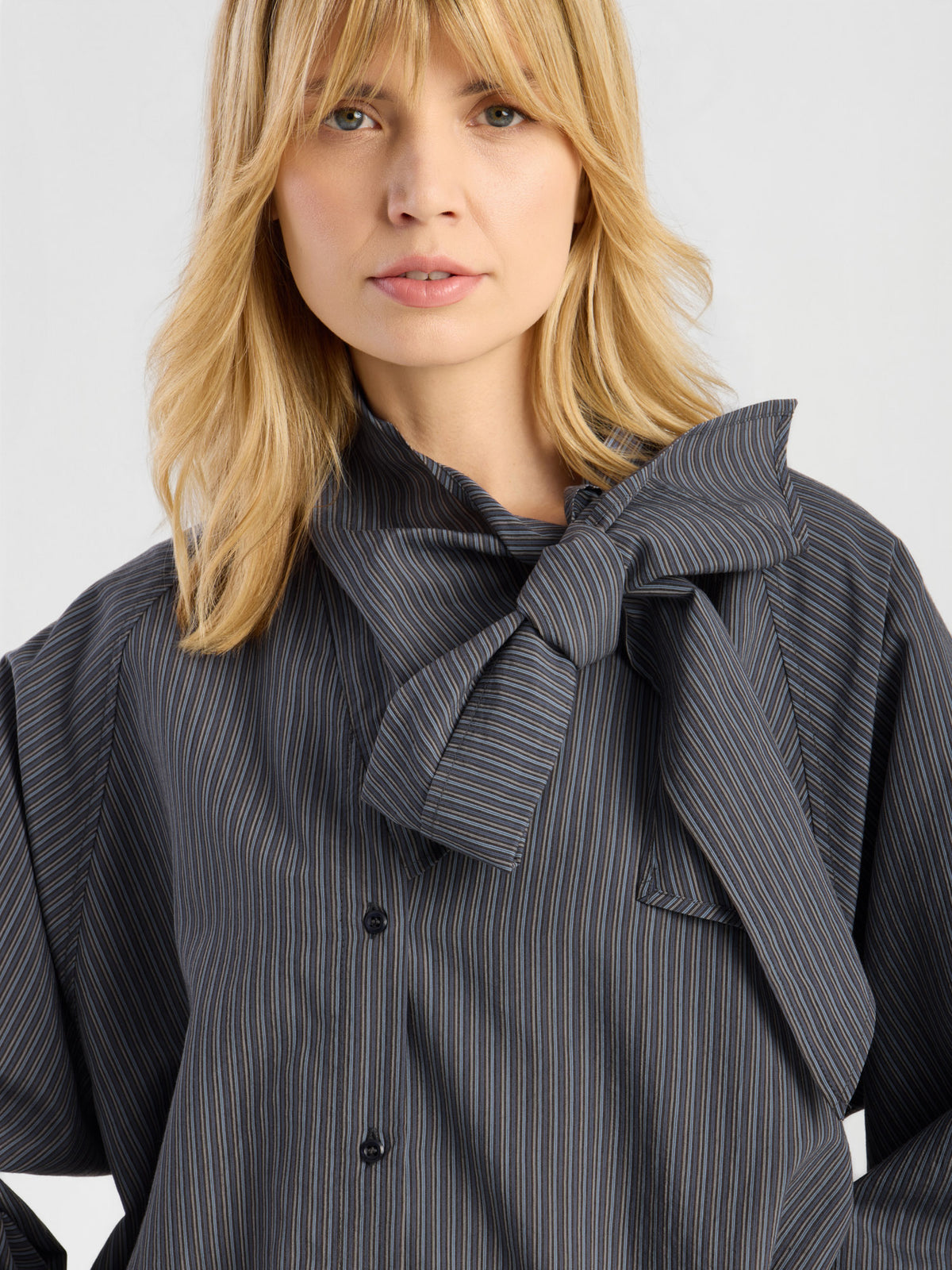 Eine Frau mit blonden Haaren trägt die „Bluse Scarf Stripes“ – eine elegante Damenbluse mit Nadelstreifen und einer auffälligen Schleife am Kragen – und steht vor einem einfarbigen Hintergrund. Hergestellt in Italien.