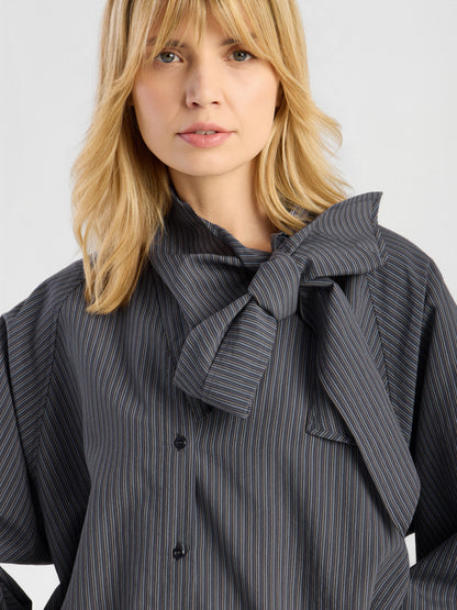 Eine Frau mit blonden Haaren trägt die „Bluse Scarf Stripes“ – eine elegante Damenbluse mit Nadelstreifen und einer auffälligen Schleife am Kragen – und steht vor einem einfarbigen Hintergrund. Hergestellt in Italien.
