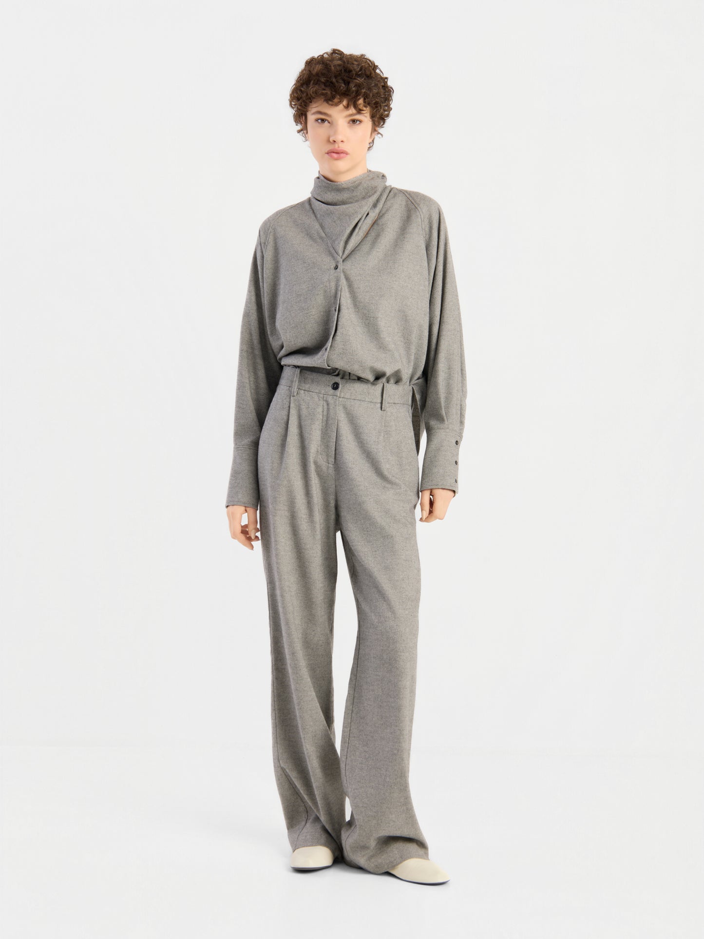 Die Person trägt den „Bluse Scarf Tweed“, ein locker sitzendes, hellgraues Rollkragenoberteil, dazu eine passende Hose mit weitem Bein und weiße Schuhe, und steht vor einem schlichten weißen Hintergrund.
