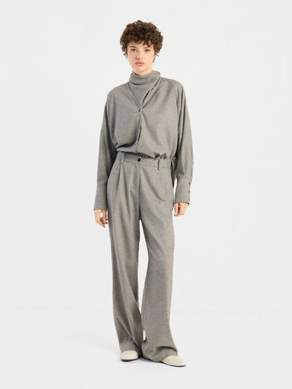 Die Person trägt den „Bluse Scarf Tweed“, ein locker sitzendes, hellgraues Rollkragenoberteil, dazu eine passende Hose mit weitem Bein und weiße Schuhe, und steht vor einem schlichten weißen Hintergrund.
