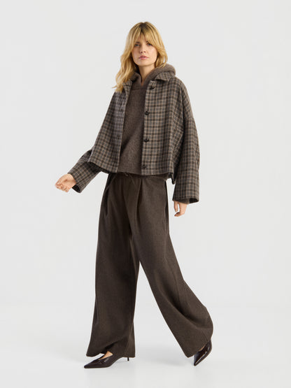 Eine Person steht vor einem schlichten Hintergrund und trägt eine weite braune Hose, ein braunes Oberteil und die kurze karierte Jacke „Barney“ mit großen Knöpfen. Sie hat blondes Haar und trägt spitze braune Schuhe.
