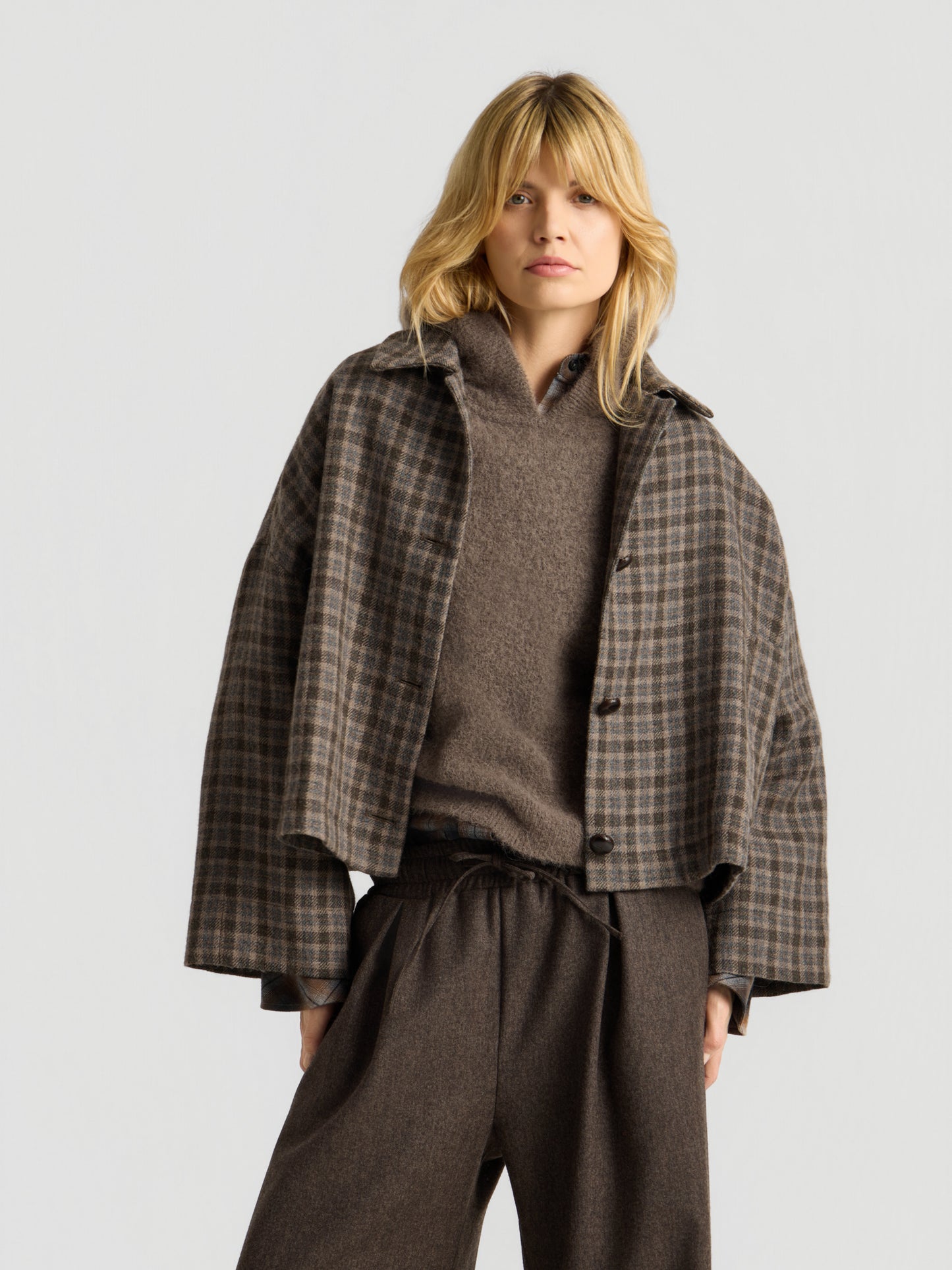 Eine Frau mit blonden Haaren trägt eine braune Karojacke über dem „Bear Hoodie Braun“ und eine weite braune Hose und steht vor einem schlichten, hellen Hintergrund.