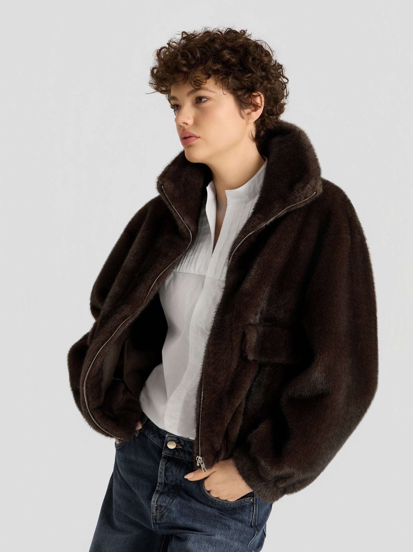 Eine Person mit kurzen Locken trägt die „Faux Mink Bomber Braun“ über einer weißen Bluse und blauen Jeans und steht vor einem schlichten hellgrauen Hintergrund.