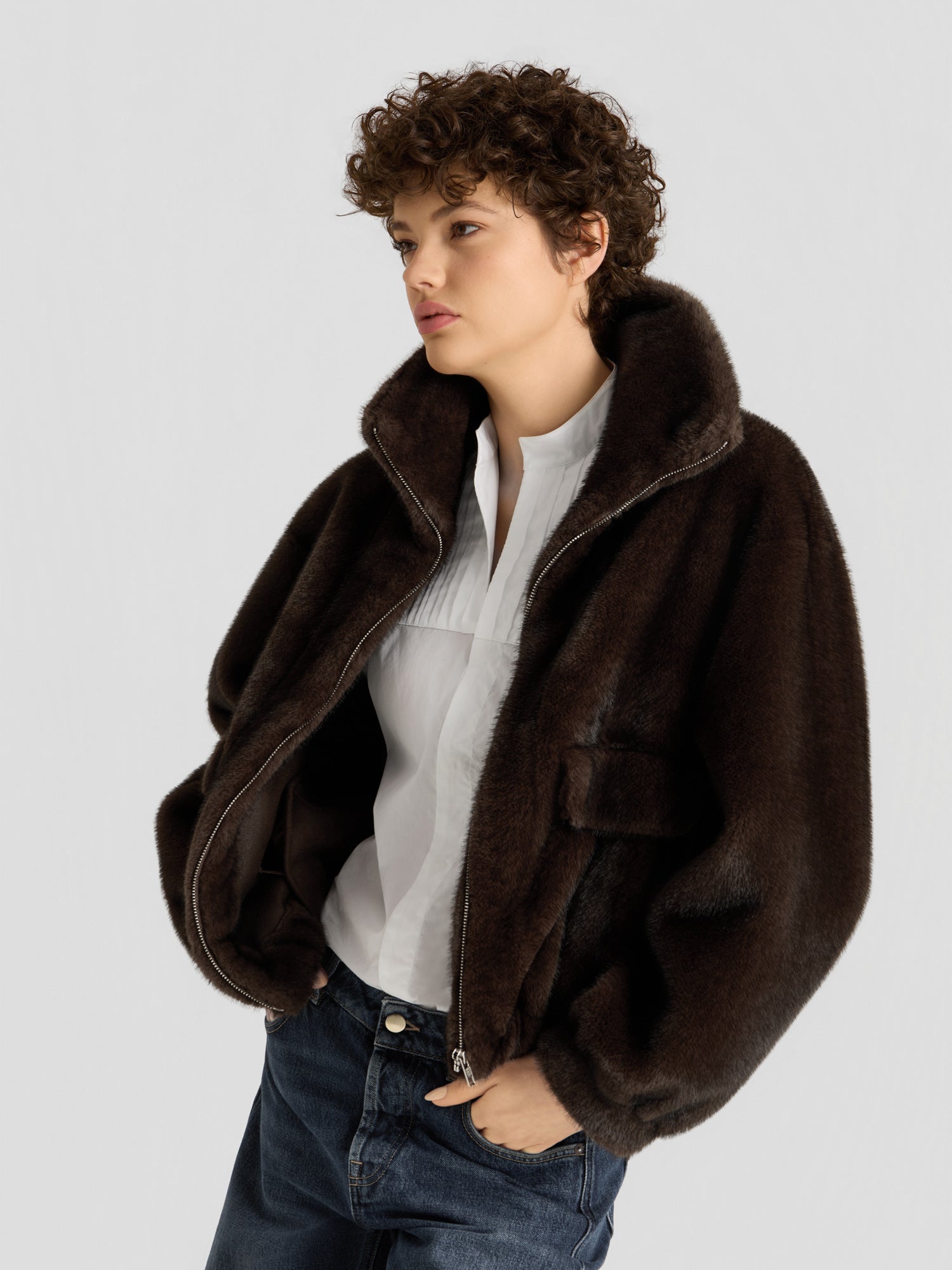 Eine Person mit kurzen Locken trägt die „Faux Mink Bomber Braun“ über einer weißen Bluse und blauen Jeans und steht vor einem schlichten hellgrauen Hintergrund.