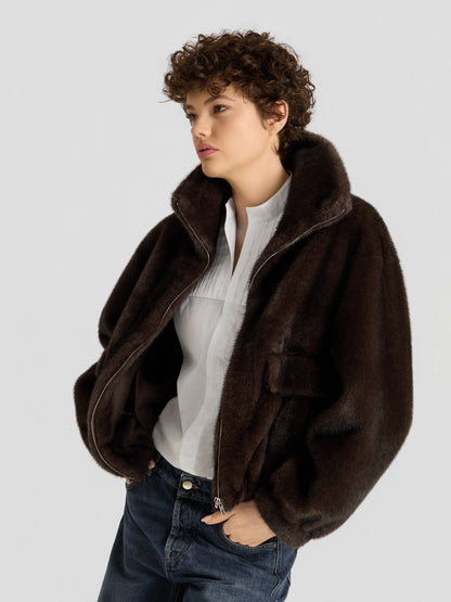 Eine Person mit kurzen Locken trägt die „Faux Mink Bomber Braun“ über einer weißen Bluse und blauen Jeans und steht vor einem schlichten hellgrauen Hintergrund.