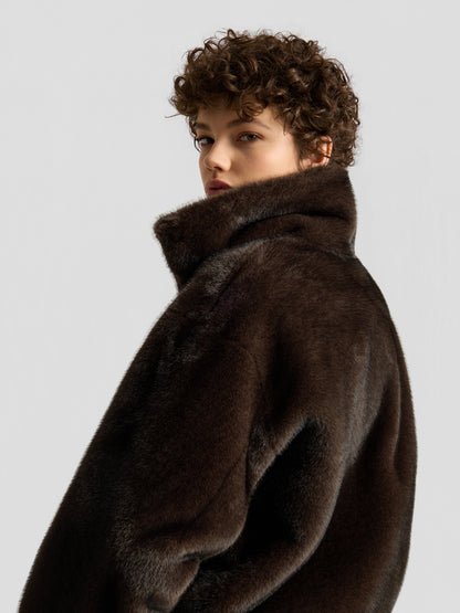 Eine Person mit kurzen Locken blickt über die Schulter; sie trägt die „Faux Mink Bomber Braun“ mit hohem Kragen vor einem schlichten, hellen Hintergrund.