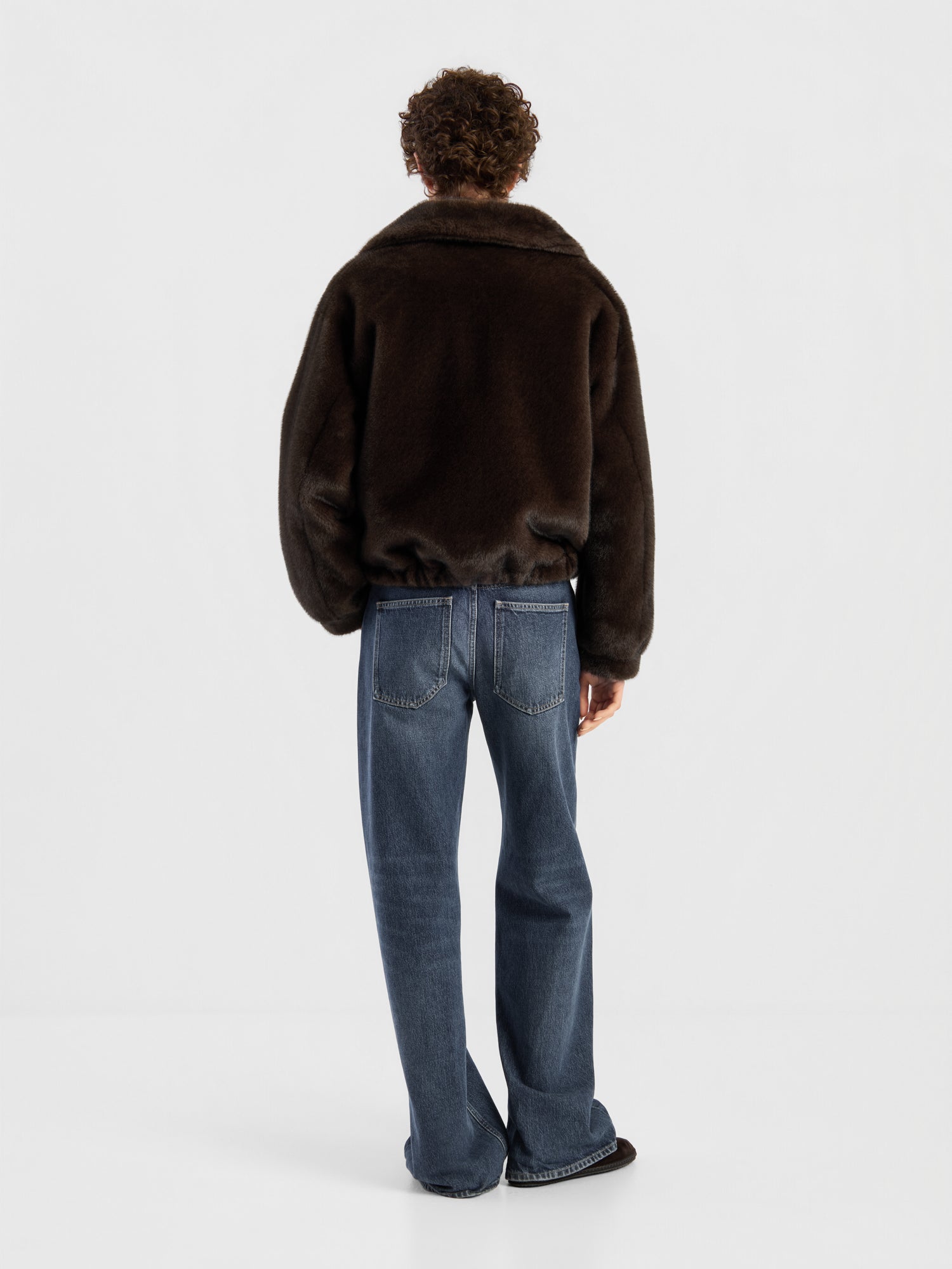 Eine Person mit lockigem Haar, die mit dem Rücken zur Kamera steht, trägt die Bomberjacke „Faux Mink“ von Braun, eine blaue Jeans und schwarze Schuhe und steht vor einem schlichten weißen Hintergrund.