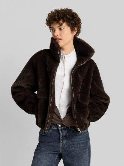 Eine Person mit kurzen Locken trägt die „Faux Mink Bomber Braun“ über einem weißen Hemd und Jeans und steht mit den Händen in den Taschen vor einem schlichten hellgrauen Hintergrund.