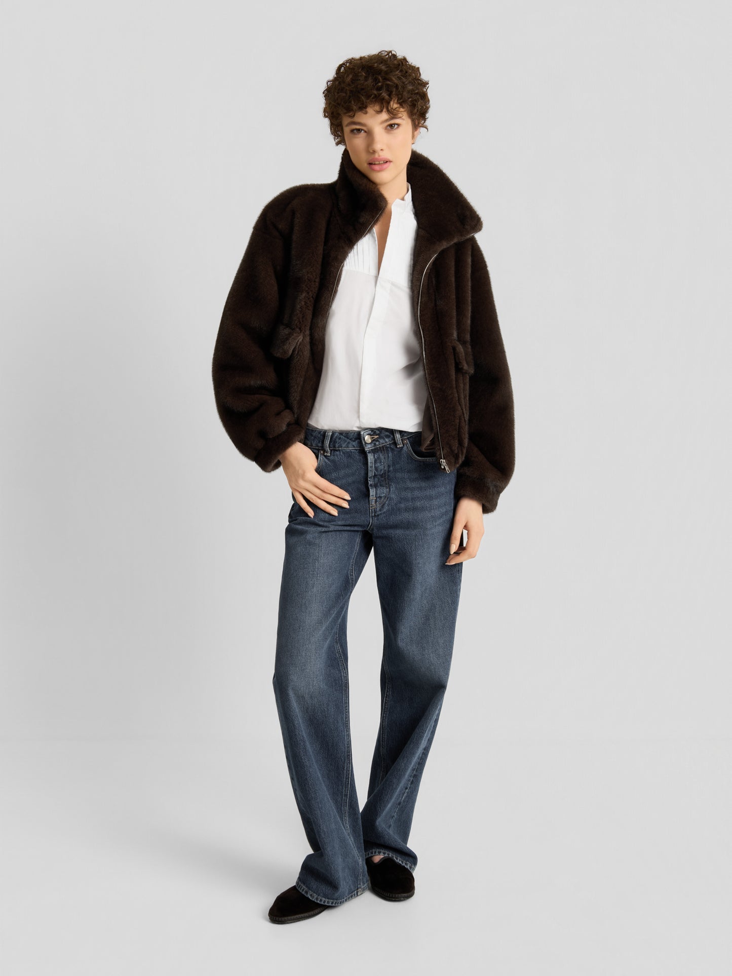 Eine Person, die die Bomberjacke „Faux Mink Bomber Braun“ trägt, kombiniert mit einer weißen Bluse, einer blauen Jeans mit weitem Bein und schwarzen Schuhen, steht vor einem schlichten hellgrauen Hintergrund.