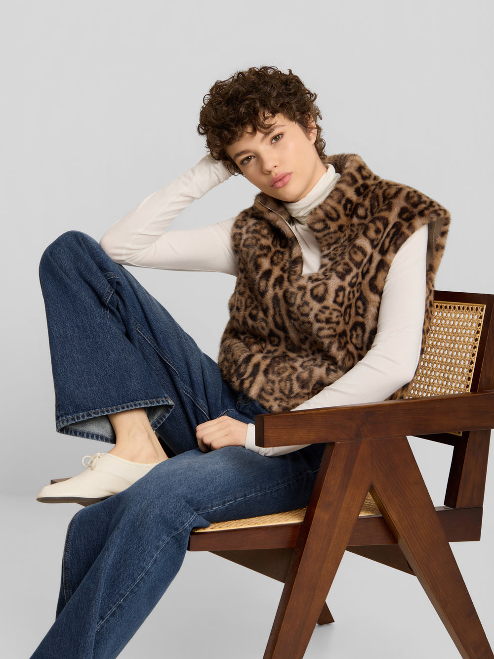 Eine Person, die eine Weste mit Leopardenmuster von A Touch of Wild über einem weißen Langarmhemd und einer blauen Jeans trägt und auf einem Holzstuhl sitzt.