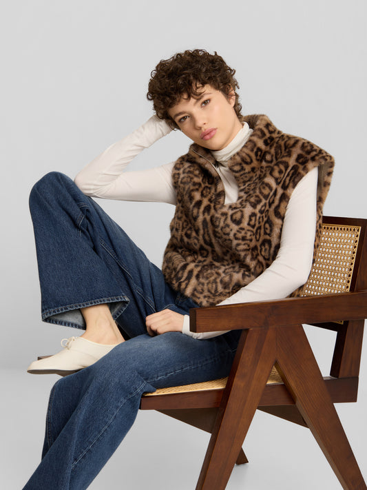 Eine Person, die eine Weste mit Leopardenmuster von A Touch of Wild über einem weißen Langarmhemd und einer blauen Jeans trägt und auf einem Holzstuhl sitzt.