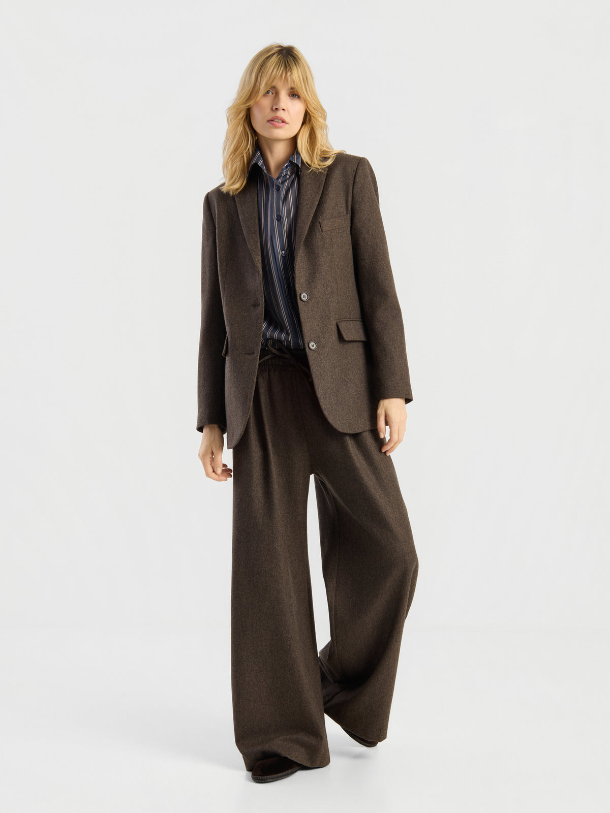 Eine Person mit blonden Haaren trägt einen braunen Oversize-Anzug mit der weit geschnittenen Hose „Pants Josy Braun“, kombiniert mit einem dunkel gestreiften Hemd, und steht vor einem schlichten weißen Hintergrund. Das Outfit strahlt lässigen Stil aus und wurde in Portugal hergestellt.