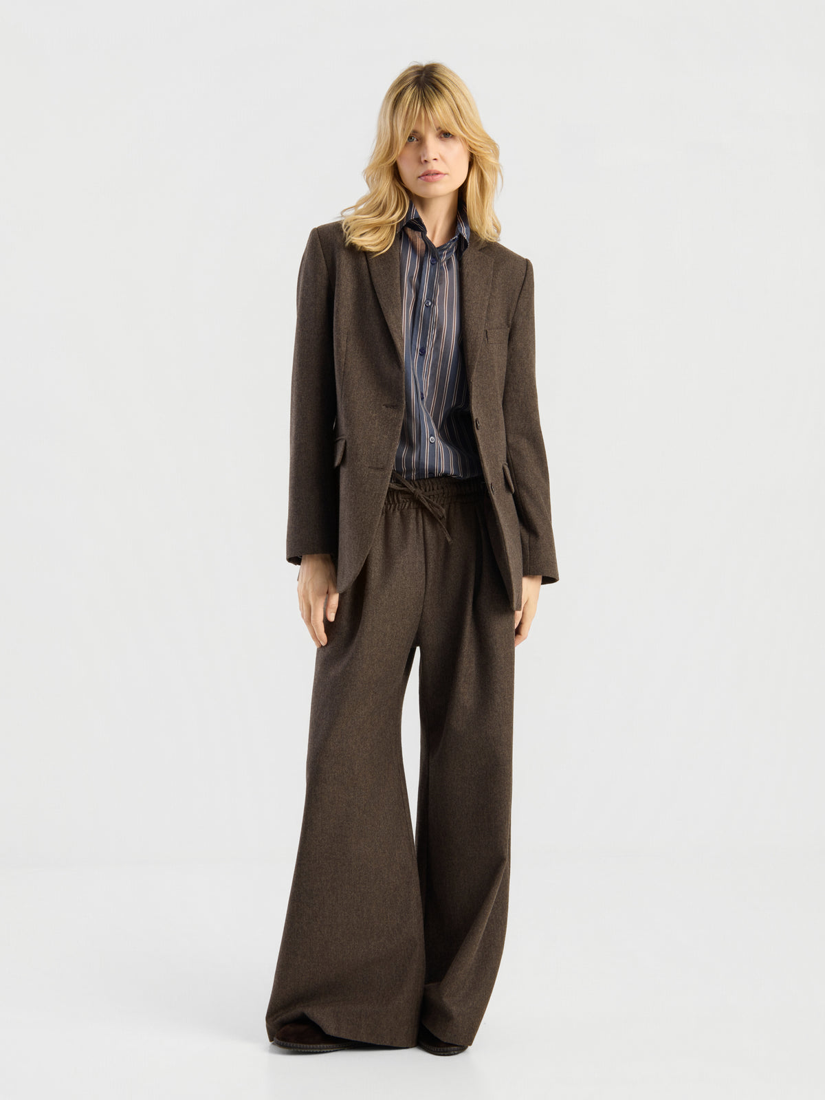 Eine Frau mit blonden Haaren trägt einen locker sitzenden Blazer von Karoline Braun, kombiniert mit einer Hose mit weitem Bein und einem gestreiften Hemd, und steht vor einem schlichten weißen Hintergrund. Dieses stilvolle Ensemble, hergestellt in Portugal, strahlt mühelose Eleganz aus.