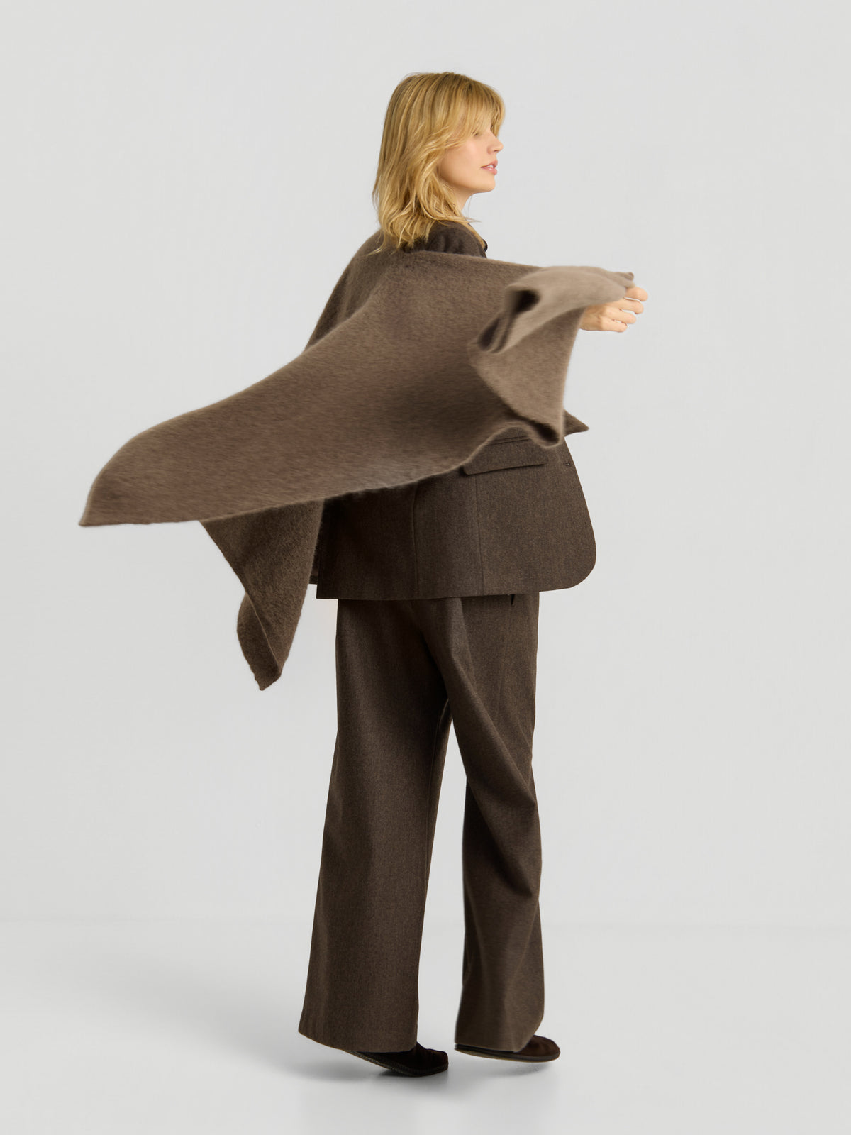 Eine Frau in einem braunen Kostüm steht seitlich im Bild und schwingt einen braunen Schal um ihre Schultern. Sie hat schulterlanges blondes Haar und posiert vor einem schlichten, hellen Hintergrund, wodurch der „Blazer Karoline Braun“ elegant zur Geltung kommt, der den zeitlosen Charme eines braunen Damenblazers unterstreicht.