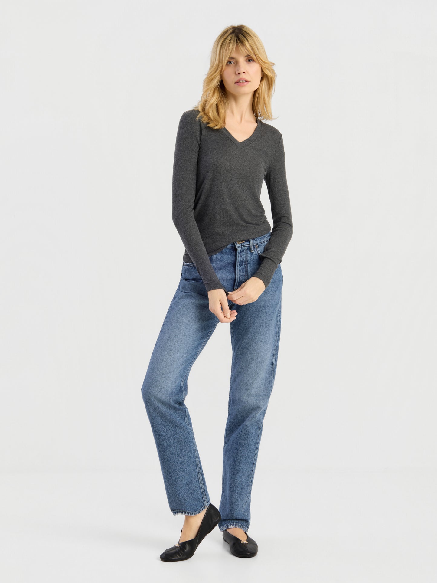 Eine Frau mit blonden Haaren trägt ein dunkles, langärmeliges Kaschmir-Shirt mit V-Ausschnitt und Rippbündchen, eine blaue Jeans und schwarze Ballerinas, während sie vor einem schlichten weißen Hintergrund steht. Sie posiert mit leicht überkreuzten Beinen und den Händen auf der Jeans. Dieses zeitlose Kleidungsstück wurde in Portugal meisterhaft gefertigt.