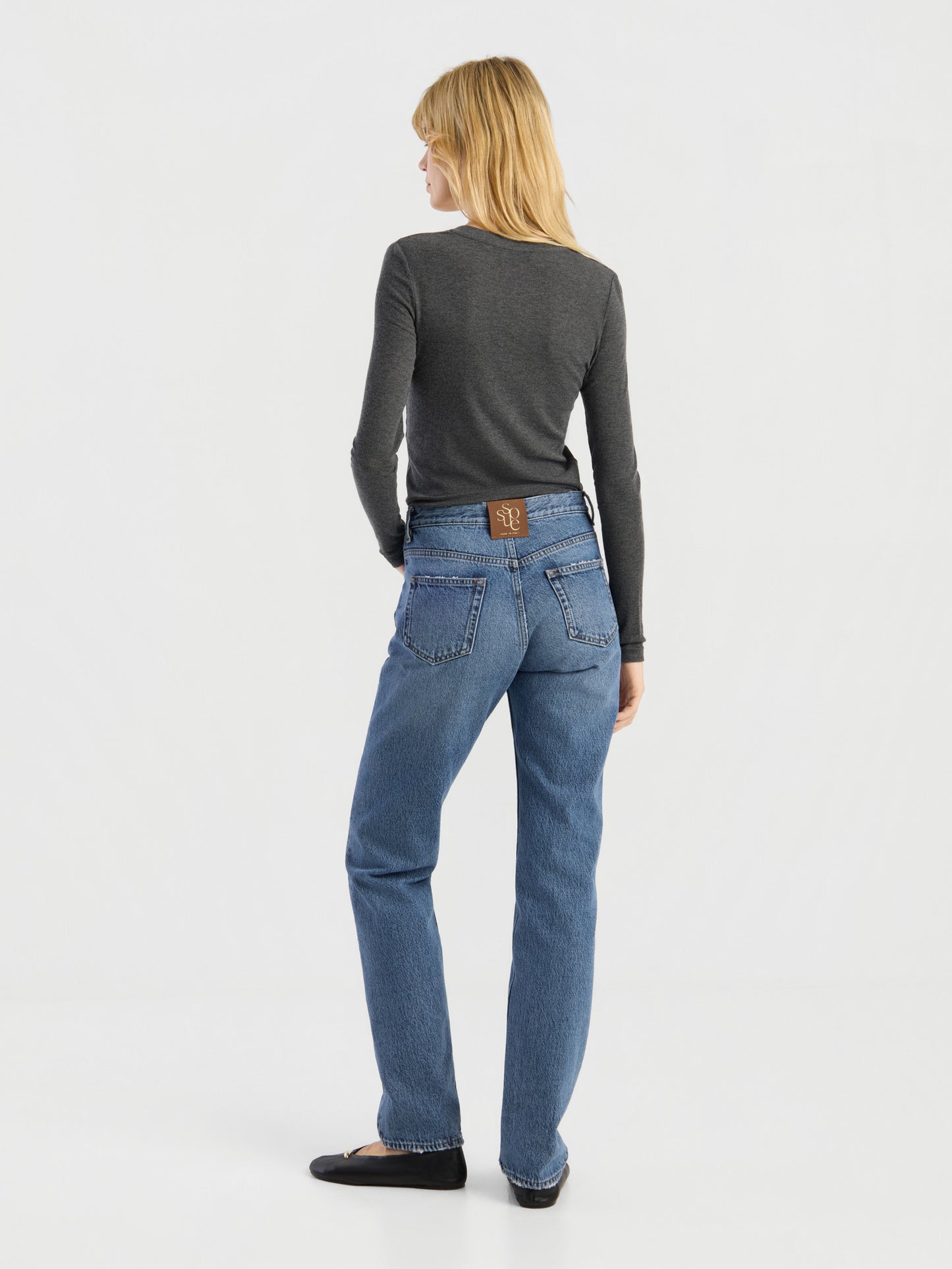 Eine Person mit schulterlangen blonden Haaren, die ein dunkelgraues Kaschmir-Langarmshirt mit V-Ausschnitt und Rippbündchen, eine Bluejeans und schwarze Schuhe trägt, steht mit dem Rücken zur Kamera in einem schlichten weißen Studio. Dieses hochwertige Shirt wird in Portugal hergestellt.