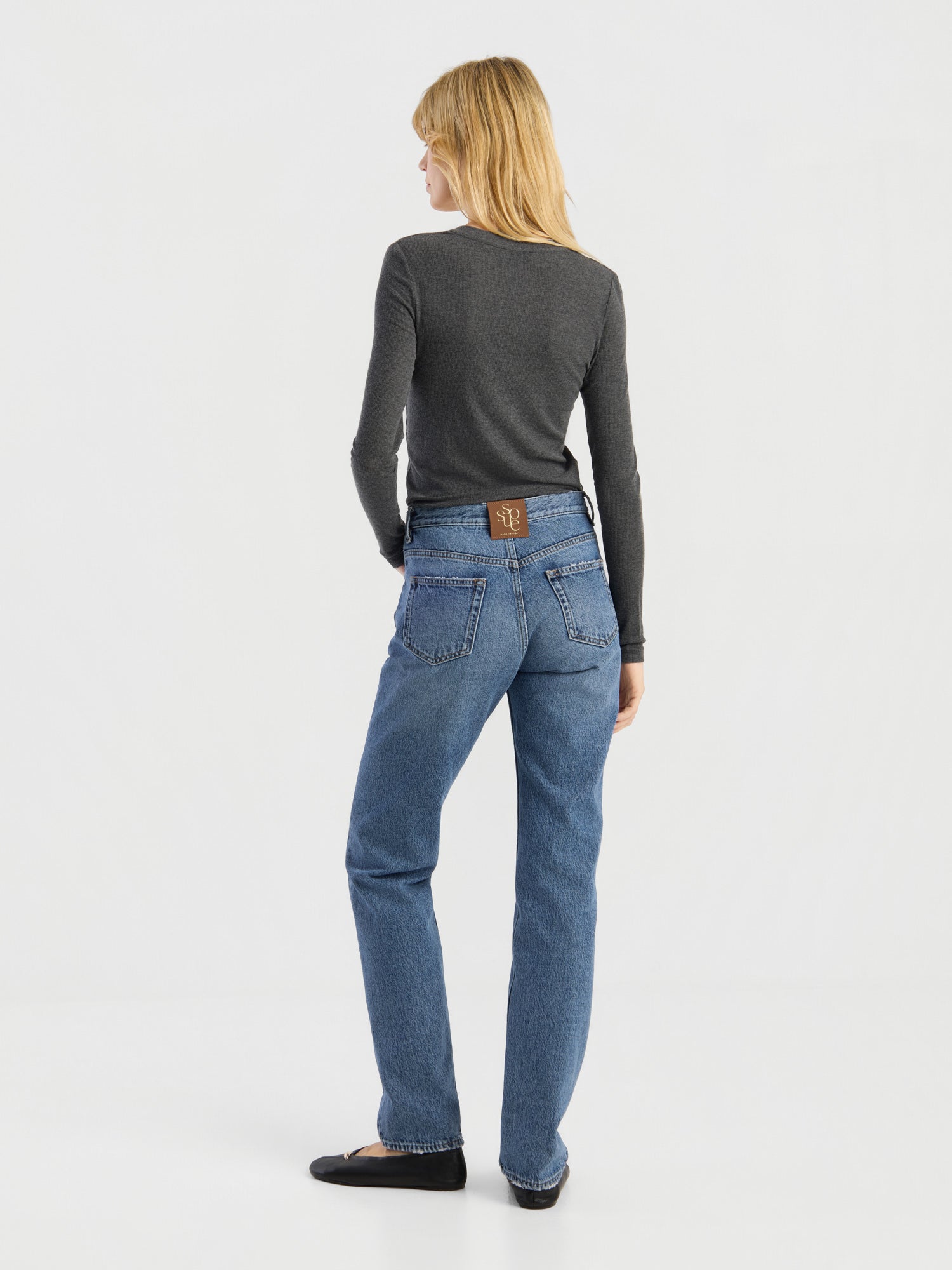 Eine Person mit schulterlangen blonden Haaren, die ein dunkelgraues Kaschmir-Langarmshirt mit V-Ausschnitt und Rippbündchen, eine Bluejeans und schwarze Schuhe trägt, steht mit dem Rücken zur Kamera in einem schlichten weißen Studio. Dieses hochwertige Shirt wird in Portugal hergestellt.