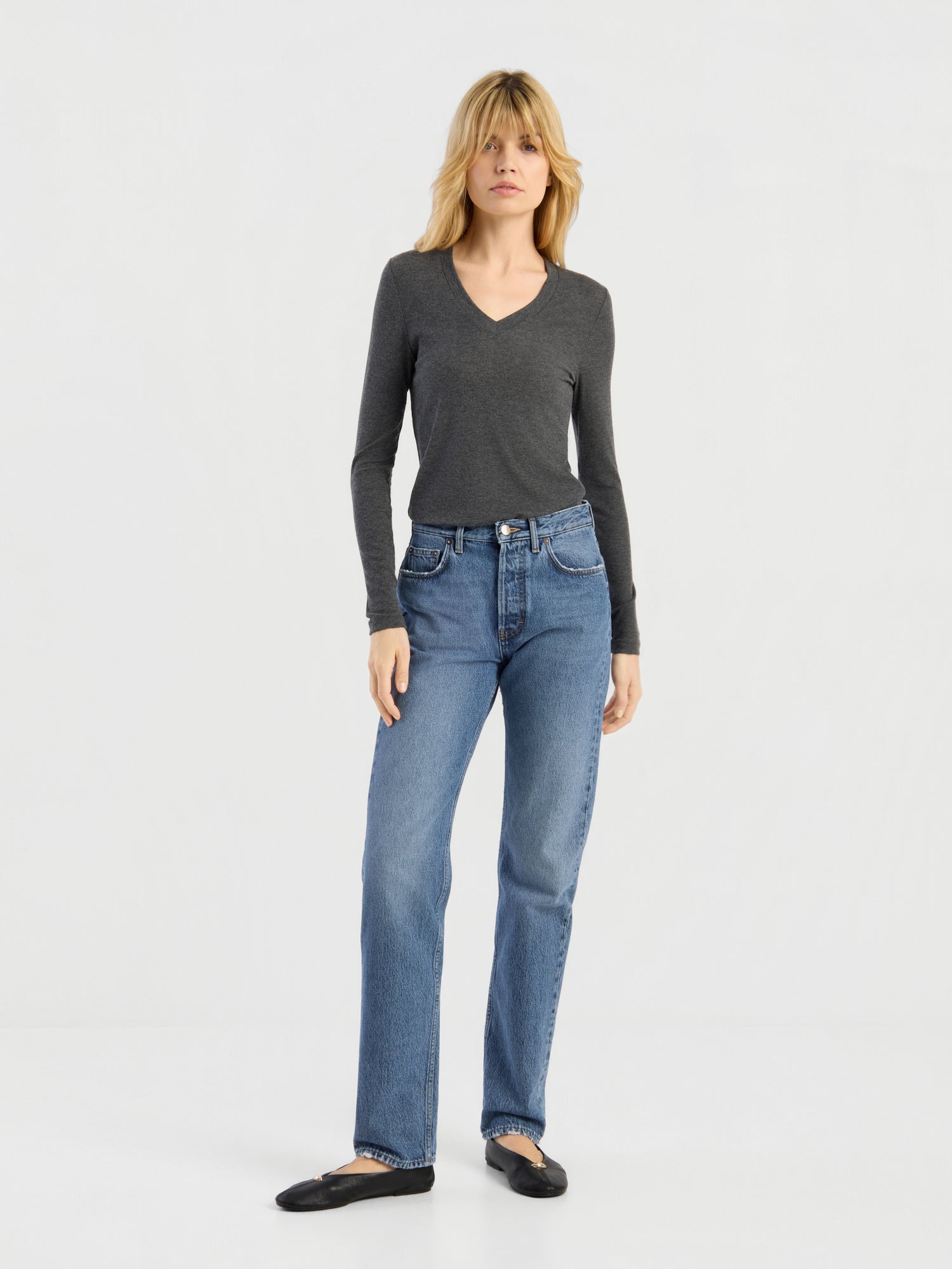 Eine Frau, die ein dunkelgraues, langärmeliges Kaschmir-Oberteil mit V-Ausschnitt und Rippbündchen, eine blaue Jeans mit geradem Bein und schwarze Ballerinas trägt, steht vor einem schlichten weißen Hintergrund. Dieses elegante Oberteil wurde in Portugal meisterhaft gefertigt.