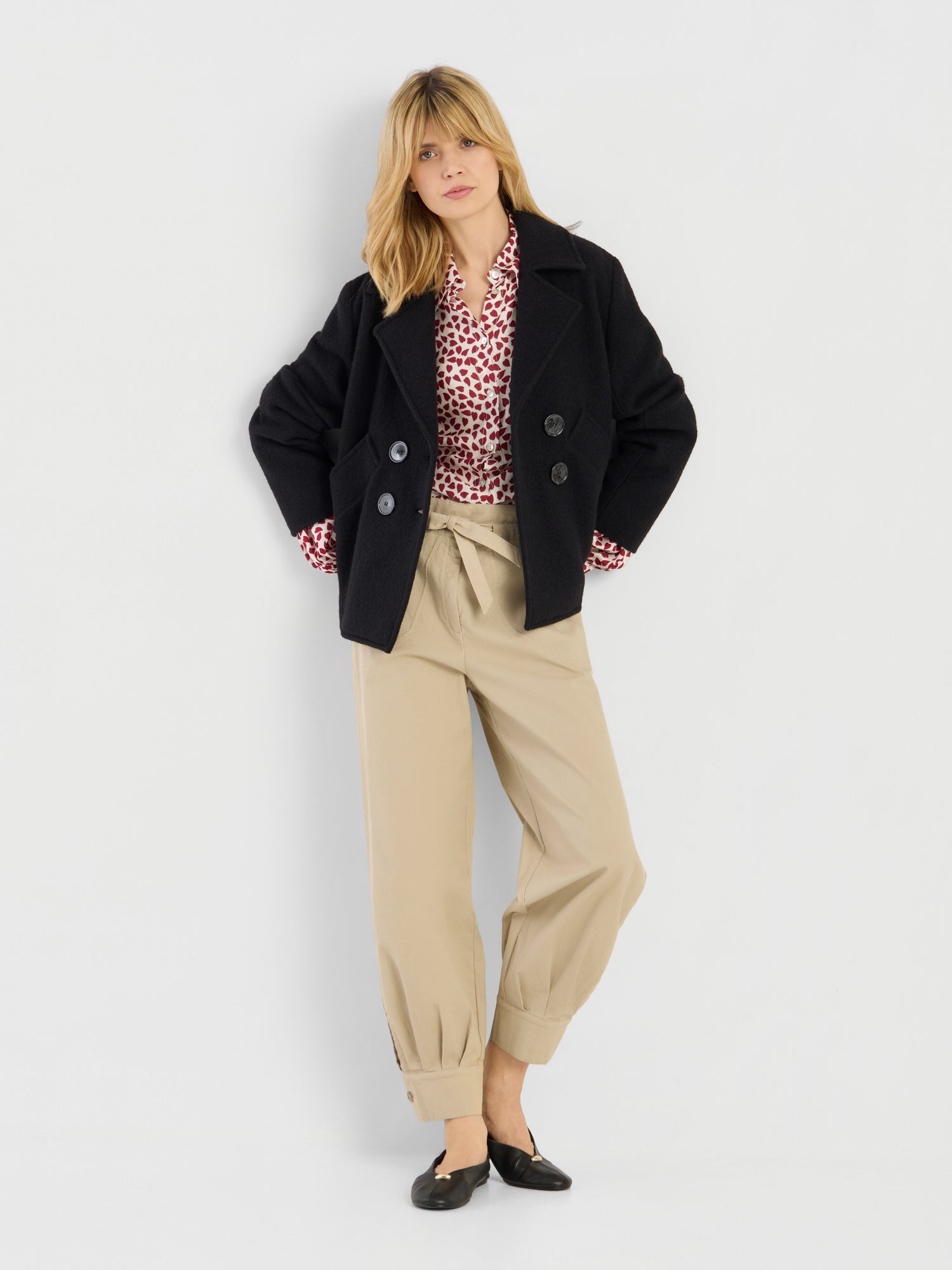 Eine Frau mit blonden Haaren posiert vor einem weißen Hintergrund. Sie trägt die hoch taillierte Hose „Love Pants Beige“ mit Umschlag, dazu einen schwarzen Peacoat, eine rot-weiß gemusterte Bluse und schwarze Schuhe.