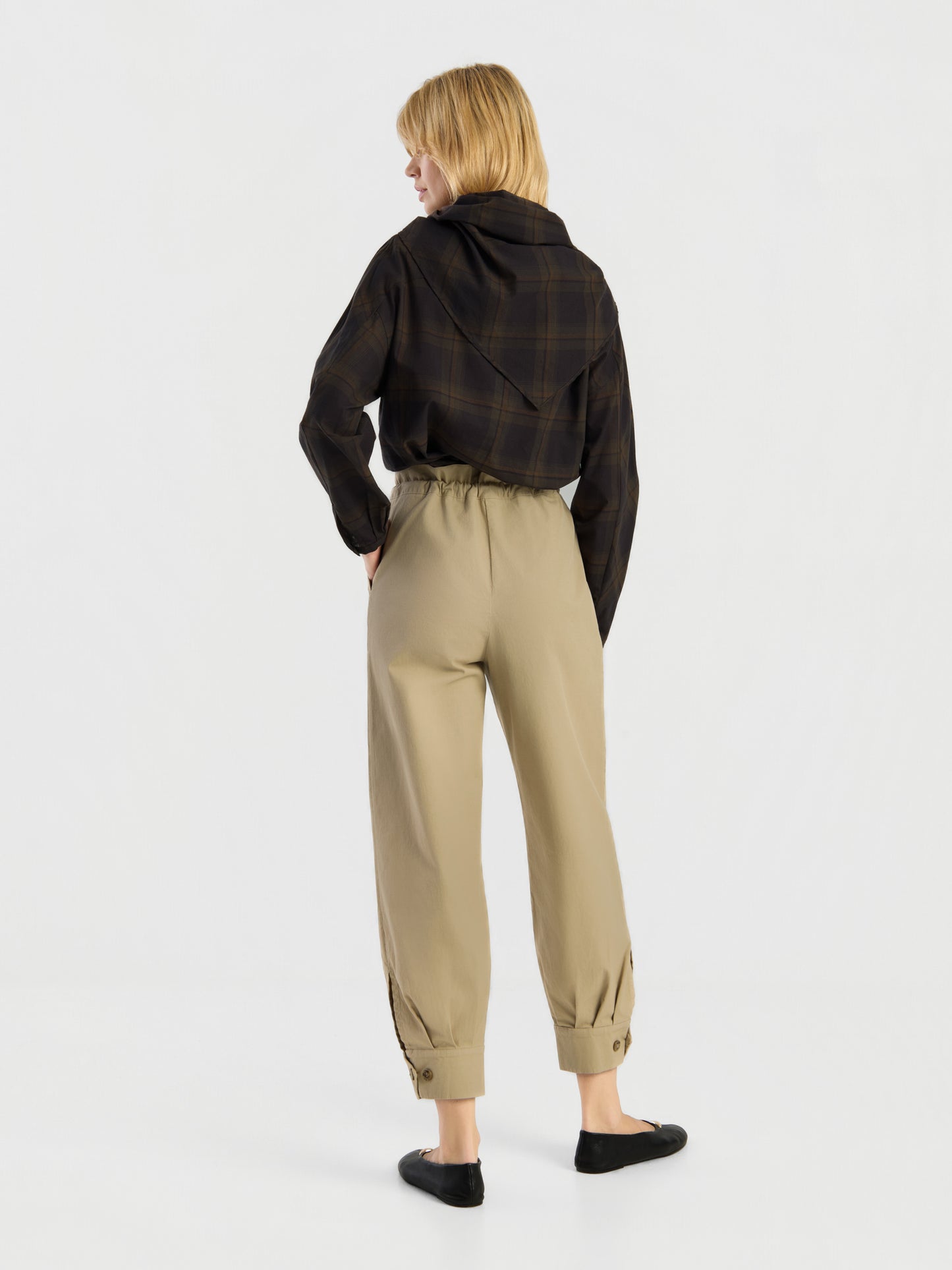 Eine Person steht mit dem Rücken zur Kamera vor einem schlichten weißen Hintergrund und trägt eine hoch taillierte „Love Pants“ in Beige mit geknöpften Bündchen, ein schwarzes Karohemd und schwarze Schuhe.