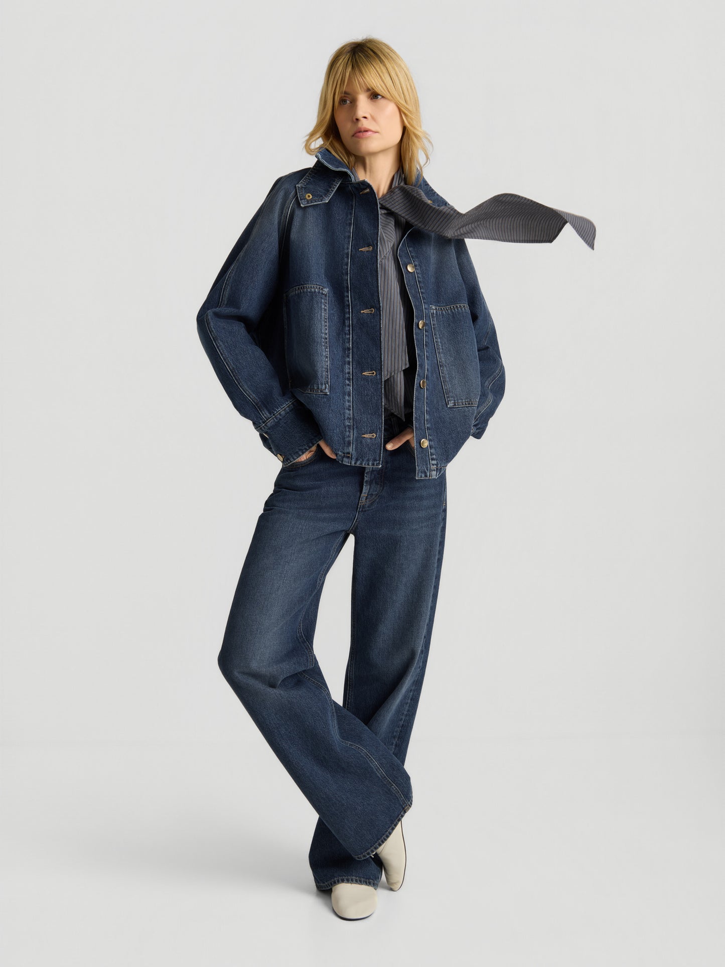 Vor einem schlichten, hellen Hintergrund steht eine blonde Person mit gekreuzten Beinen und trägt eine dunkelblaue Oversized-Denim-Bomberjacke, eine Jeans mit weitem Bein, ein dunkles Hemd, weiße Schuhe und einen gestreiften Schal, der zur Seite weht.