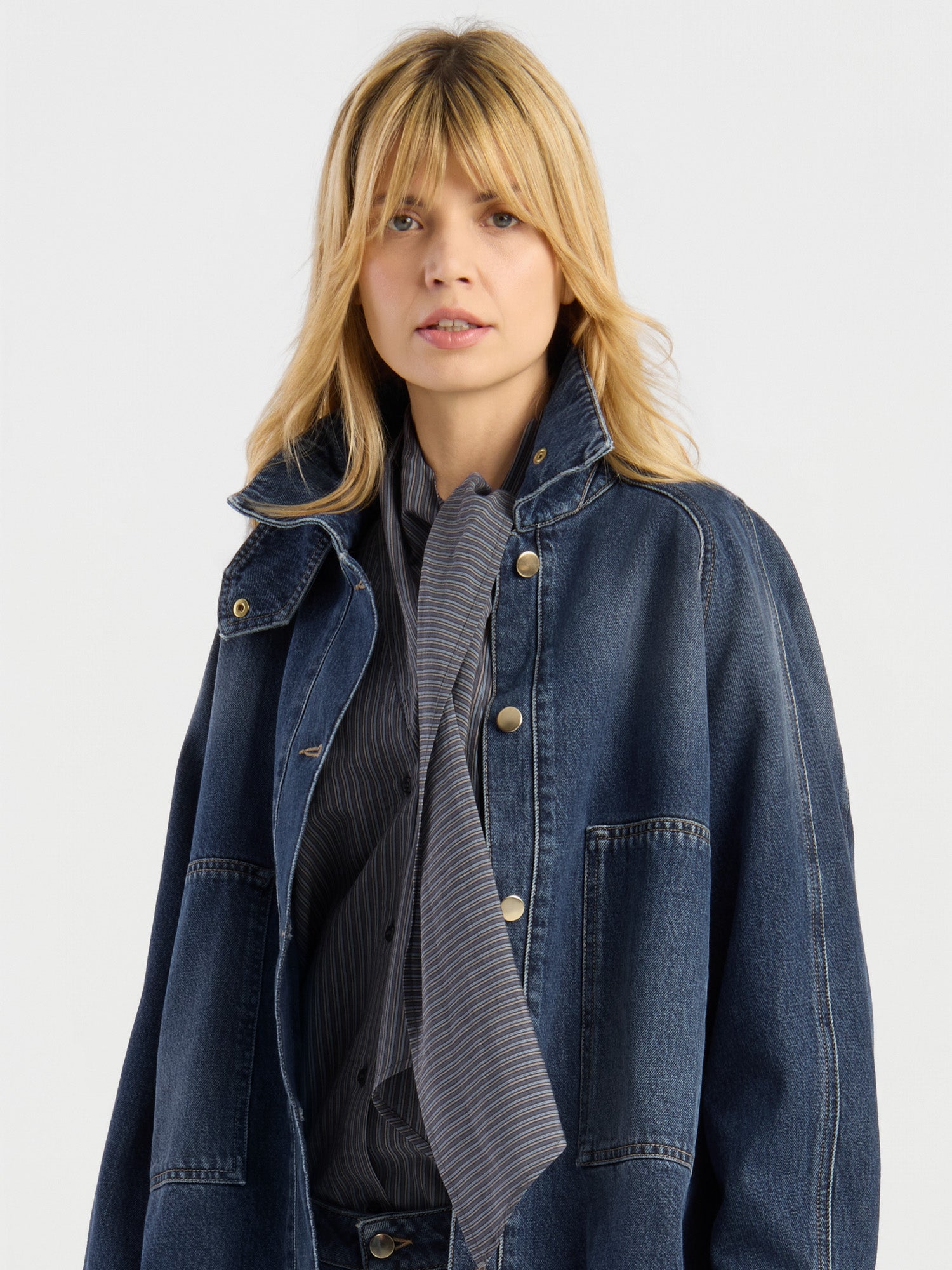Eine blonde Frau, die die Oversized Denim Bomber Dark Blue über einem dunkel gestreiften Hemd und einem dazu passenden Schal trägt, steht vor einem schlichten hellen Hintergrund.