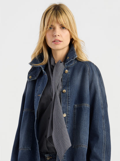 Eine blonde Frau, die die Oversized Denim Bomber Dark Blue über einem dunkel gestreiften Hemd und einem dazu passenden Schal trägt, steht vor einem schlichten hellen Hintergrund.