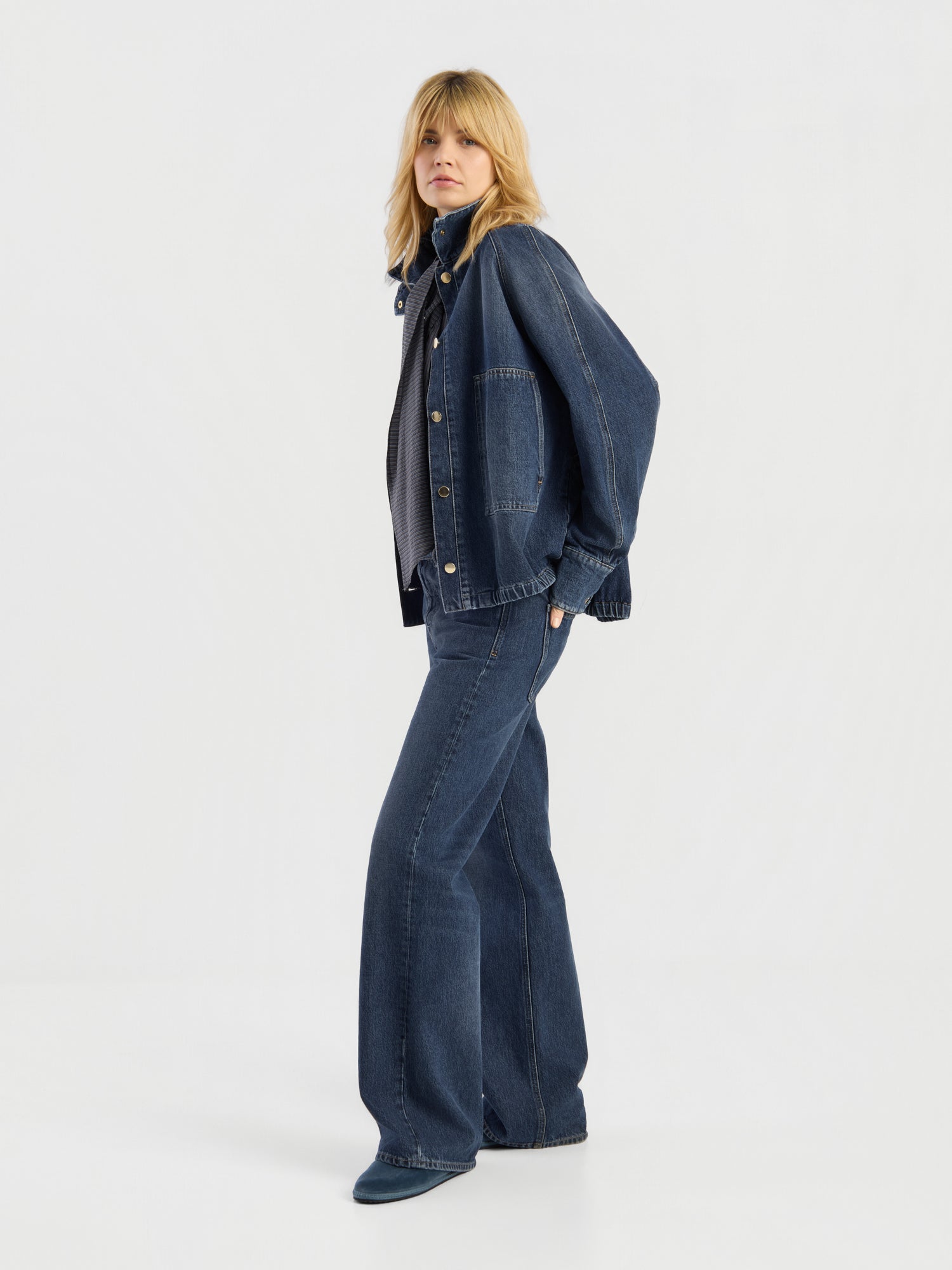 Eine Person mit blonden Haaren steht im Profil vor einem schlichten, hellen Hintergrund und trägt die „Oversized Denim Bomber Dark Blue“, ausgestellte Jeans und Freizeitschuhe.