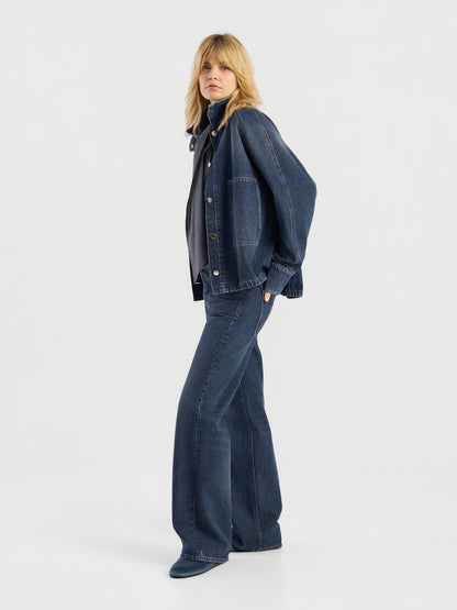 Eine Person mit blonden Haaren steht im Profil vor einem schlichten, hellen Hintergrund und trägt die „Oversized Denim Bomber Dark Blue“, ausgestellte Jeans und Freizeitschuhe.