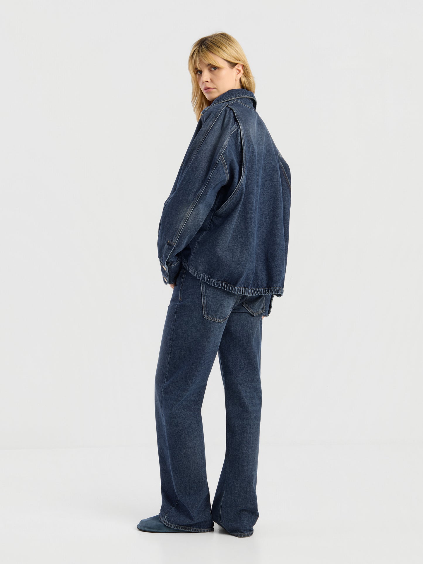 Eine Person mit blonden Haaren trägt die Oversized Denim Bomber Dark Blue und dazu passende Jeans mit weitem Bein, steht vor einem schlichten weißen Hintergrund und blickt über die Schulter.
