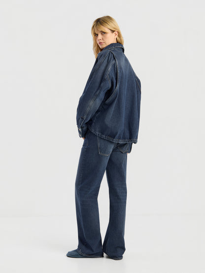 Eine Person mit blonden Haaren trägt die Oversized Denim Bomber Dark Blue und dazu passende Jeans mit weitem Bein, steht vor einem schlichten weißen Hintergrund und blickt über die Schulter.